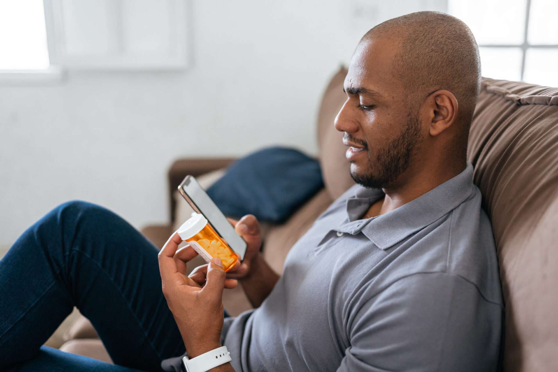 man on couch reading pill bottle and checking smartphone